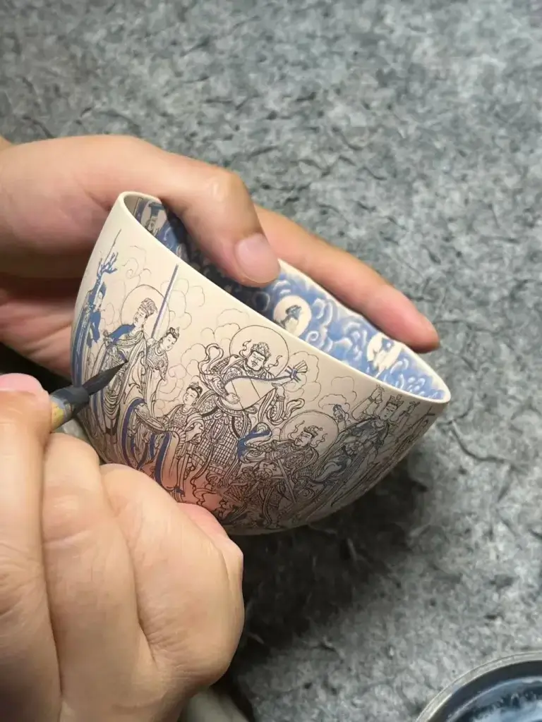Chinese artisan hand-painting intricate deity figures on a raw porcelain teacup, demonstrating traditional Jingdezhen ceramic craft